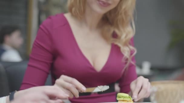 Retrato bela mulher loira com cabelo encaracolado comer bolo com um macaroon no restaurante de perto. Senhora quente com decote profundo goza de sua sobremesa. Câmara move-se — Vídeo de Stock
