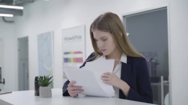 Portre resmi giysiler içinde düşünceli genç bayan dikkatle tezgahta duran ofiste kağıtları kontrol ilgili. Uzun saçlı kadın problem çözme. Çalışma süreci. Kamera sola hareket eder