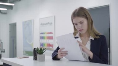 Resmi kıyafetler içinde endişeli düşünceli genç bayan özenle tezgahın başında duran ofiste kağıtları kontrol. Uzun saçlı kadın üzgün, o sorunu çözmeye çalışıyor. Kamera sağa doğru hareket eder