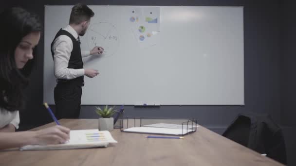 Young man drawing pie chart on the office board showing information to female colleague involved in work. Brunette woman drawing in her notebook sitting at the table, looking on the board. Workplace — ストック動画