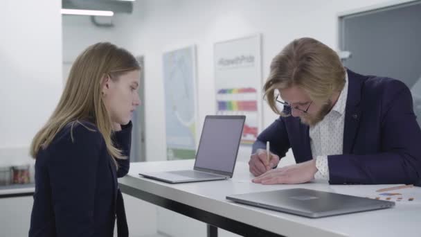 Jonge man met een baard met een bril die informatie op het papier wikkelde terwijl zijn vrouwelijke collega naar hem stond te kijken aan de toonbank in het kantoor. Werkmomenten — Stockvideo