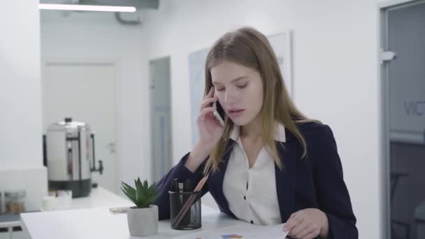 카운터에 서있는 사무실에서 휴대 전화 확인 서류로 말하는 정장 드레스를 입은 젊은 아가씨. 문제를 해결 긴 머리를 가진 여자. 직장. 사무실 공간. — 비디오