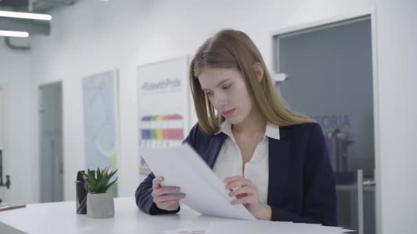 Jovencita reflexiva vestida con ropas formales revisando atentamente los documentos en la oficina de pie en el mostrador. Mujer de pelo largo sorprendida por lo que está escrito en papel. Lugar de trabajo. Espacio de oficina . — Vídeos de Stock