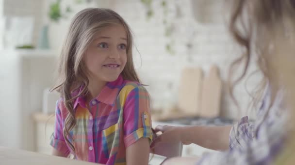 Retrato de una niña linda con hermosos ojos grandes con una camisa multicolor en el interior. Mamá endereza el cabello de sus hijas. Relaciones madres e hijas. Una familia feliz . — Vídeos de Stock