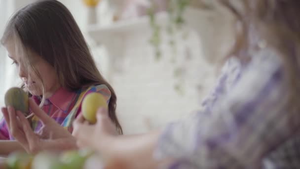 Mano femenina sosteniendo huevo de Pascua mientras pequeña niña sonriente pintando en ella un corazón con un pequeño cepillo de cerca. Preparación para las vacaciones de Pascua. Madre e hija se divierten juntas en la cocina — Vídeos de Stock