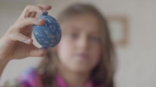 Figura borrosa de la niña sosteniendo huevo de Pascua azul con el corazón pintado en la mano, mostrándolo a la cámara. El foco está en la mano. Concepto de creatividad, tradiciones. Preparación de la Pascua . — Vídeos de Stock