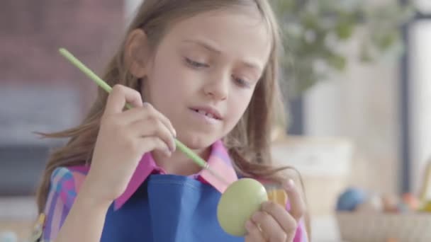 Portret mooi klein meisje met prachtig haar in blauwe schort schilderij paasei met een kleine borstel zitten in de keuken. Voorbereiding op de paasvakantie. — Stockvideo