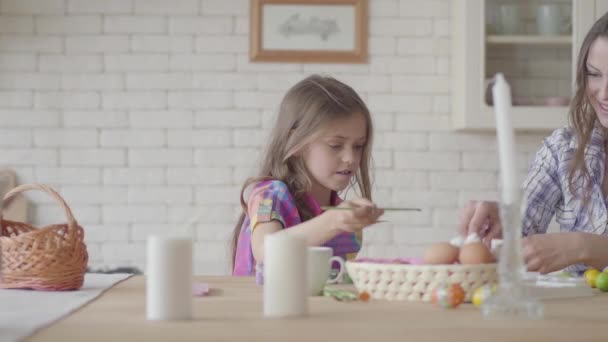 Linda niña sonriente pintando huevos orientales con un cepillo pequeño sentado en la mesa con su madre en la cocina. Preparación para las vacaciones de Pascua. Madre e hija se divierten juntas en la cocina — Vídeos de Stock