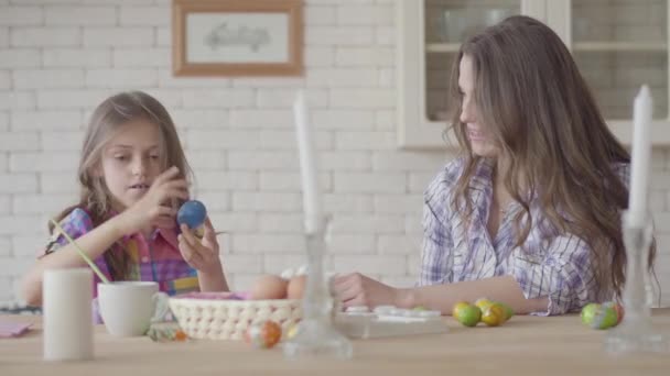 Portret van moeder en dochter zittend aan de paastafel. Schattig meisje schildert een paasei met een dunne kwast. Relatie moeders en dochters. Een gelukkige familie. Voorbereiding van het Pasen. — Stockvideo