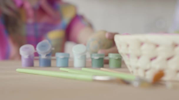 Pequeñas botellas de pintura acrílica y pinceles tumbados sobre la mesa se cierran. Las manos de la niña pusieron el pincel en la botella y pintaron el huevo de Pascua. Primer plano y fondo desenfocado — Vídeo de stock