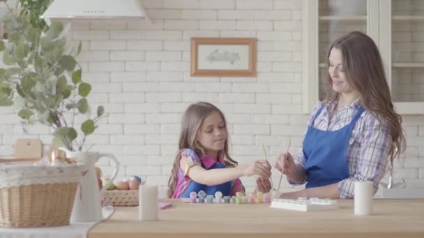 Menina segurando ovo de Páscoa e pintá-lo com sua mãe usando um pequeno pincel. Uma família feliz. Preparação da Páscoa . — Vídeo de Stock