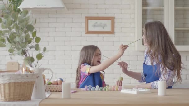Mała dziewczynka malowanie jaj z matką za pomocą małego pędzla. Dziecko próbuje dotknąć kobiety nos z pędzlem. Przygotowanie do Świąt Wielkanocnych. Matka i córka bawią się razem w kuchni — Wideo stockowe