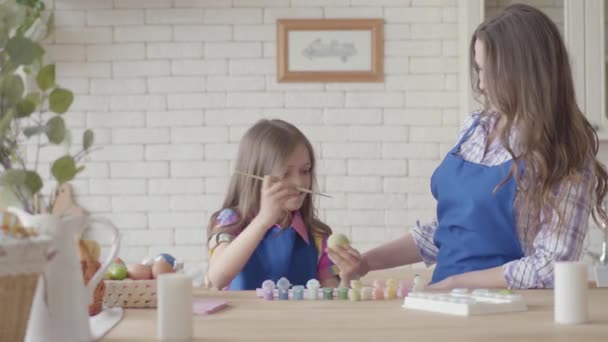 Carina ragazza con i capelli lunghi pittura uovo di Pasqua con un pennello a tavola in cucina. Bella donna in piedi vicino e sorridente. Preparazione per le vacanze di Pasqua. Concetto di creatività — Video Stock