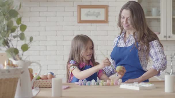 Mutfakta masada bir fırça kullanarak uzun saç boyama Paskalya yumurtası ile Portre sevimli kız. Güzel kadın yanında duruyor ve gülümsüyor. Paskalya tatili için hazırlık. Yaratıcılık kavramı — Stok video