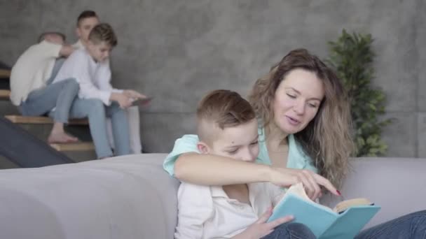 Retrato de la hermosa madre joven leyendo el libro a su hijo mientras el resto de sus hijos adolescentes jugando entre sí en el fondo sentado en las escaleras con la tableta. Gran familia ruidosa amistosa — Vídeos de Stock