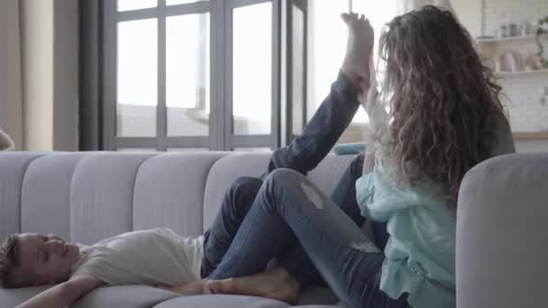 Mooie jonge moeder zittend op de Bank met haar kleine zoon. De jongen spelen met de vrouw met zijn benen, Lady neemt zijn voeten en het maken van massage. Gelukkige vriendelijke familie — Stockvideo