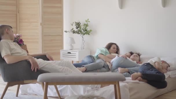 Jonge moeder met haar tienerzoons die samen slapen in het grote bed. Eén jongen zit op de stoel. Gelukkig liefdevol familieconcept. Rustig uurtje in de familie met veel kinderen — Stockvideo