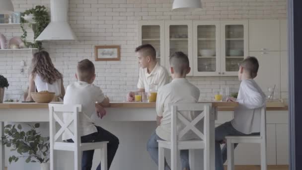 Young pretty mother bakes pies for her four teen sons in white shirts waiting for the food sitting at the table in the kitchen. Big friendly noizy family having breakfast together — 비디오