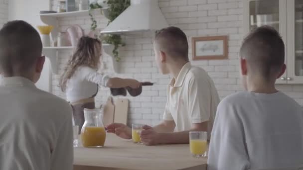 Die junge hübsche Mutter backt Kuchen für ihre vier jugendlichen Söhne in weißen Hemden, die am Tisch in der Küche auf das Essen warten. große freundliche lärmende Familie beim gemeinsamen Frühstück — Stockvideo