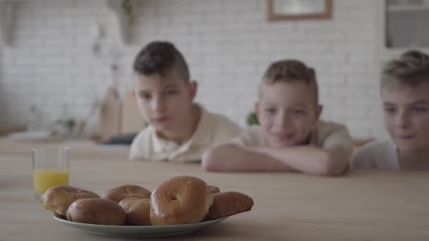 Drei Jungen schauen auf den Teller, im Hintergrund stehen leckere kleine Kuchen auf dem Tisch. Jungen haben Hunger. Konzept der Fütterung — Stockvideo