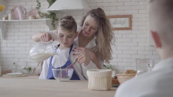 Ładna kobieta i jej nastoletni syn gotowania w kuchni, chłopiec pomaga matce zrobić jedzenie. Mama nalewanie mleka w misce i mieszanie go, nauczanie chłopca. Drugi syn patrzy na pierwszym planie — Wideo stockowe