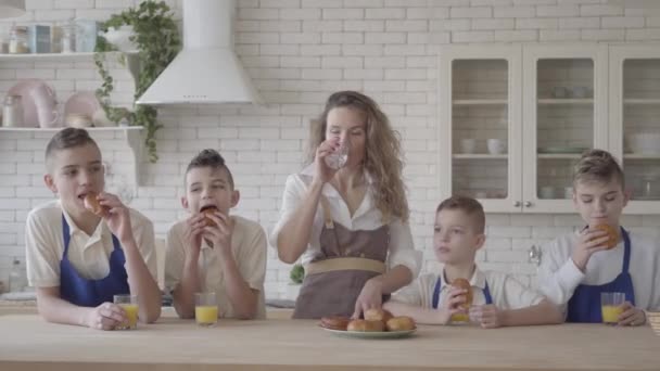 Portrait séduisante femme heureuse et ses quatre fils adolescents mangeant des tartes et buvant du jus d'orange dans le chaton debout à la table. Tout le monde porte des tabliers. Enfants aidant mère à cuisiner la nourriture — Video