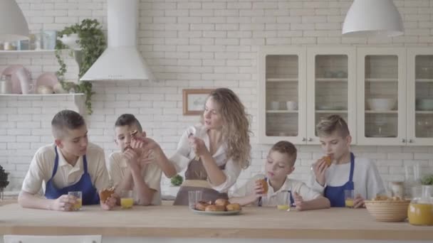 Portrait séduisante femme heureuse et ses quatre fils adolescents mangeant des tartes et buvant du jus d'orange dans le chaton debout à la table. Tout le monde porte des tabliers. Enfants aidant mère à cuisiner la nourriture — Video