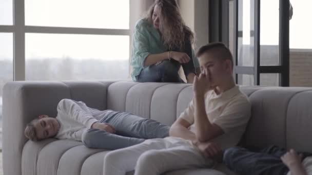 Jonge mooie moeder zit op de bank in de woonkamer met haar tienerzoons. Een vrouw die haar kinderen ziet spelen, grappen maken en kalmeren. Happy grote vrolijke liefdevolle familie — Stockvideo