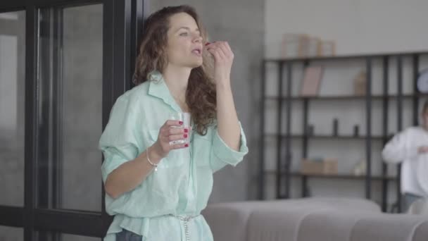 Portret moe jonge moeder proberen te kalmeren zichzelf staande in de buurt van de muur met glas water terwijl haar tienerzonen rennen araund spelen met elkaar op de achtergrond. — Stockvideo