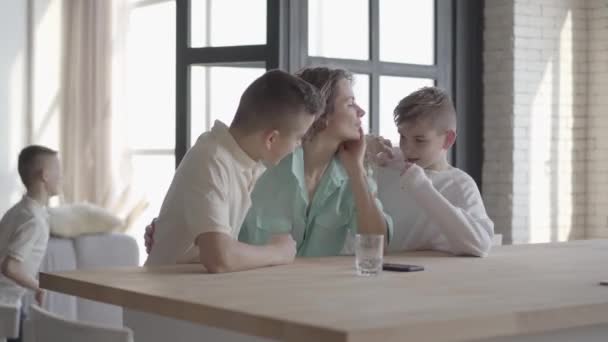 Jeune jolie femme aux cheveux bouclés debout au comptoir à la maison, ses petits fils adolescents courent partout en jouant ensemble. Grande famille amicale heureuse — Video