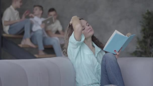Porträt einer hübschen jungen Mutter, die auf dem Sofa ein Buch liest, während ihre Teenie-Kinder auf der Treppe im Hintergrund mit dem Tablet plaudern. Ein Junge kommt und hält die Frau lächelnd in der Hand. große freundliche — Stockvideo