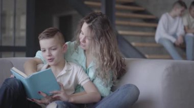 Güzel genç anne portresi oğlu kanepede otururken onun iki genç çocuklar arka planda merdivenlerde otururken kitap okuma. Büyük dostu gürültülü aile