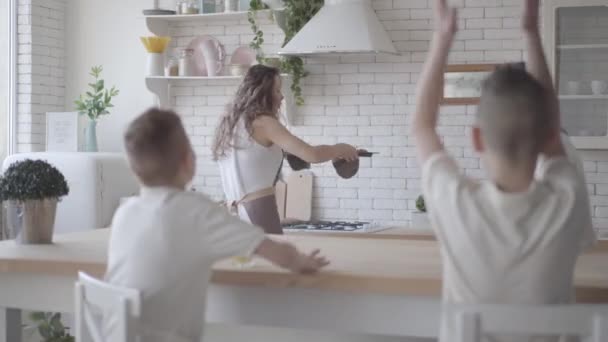 Portre genç güzel anne mutfakta masada oturan yiyecek bekliyor beyaz gömlek onu dört genç oğulları için turta pişirir. Büyük dostu gürültülü aile birlikte kahvaltı yapıyor — Stok video