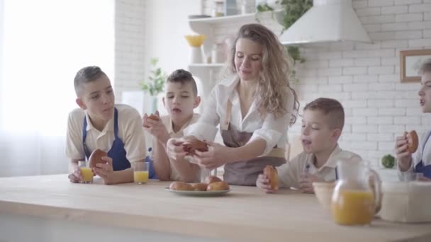 Atractiva mujer feliz y sus cuatro hijos adolescentes comiendo pasteles y bebiendo jugo de naranja en el kitcken de pie en la mesa. Todos usando delantales. Niños ayudando a la madre a cocinar alimentos — Vídeos de Stock