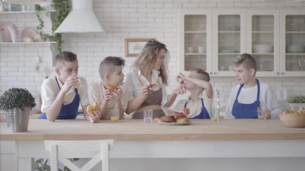 Портрет привабливої щасливої жінки та її чотирьох синів-підлітків, які їдять пироги та п'ють апельсиновий сік у кіккені, що стоїть за столом. Усі носять фартухи. Діти допомагають мамі готувати їжу — стокове відео