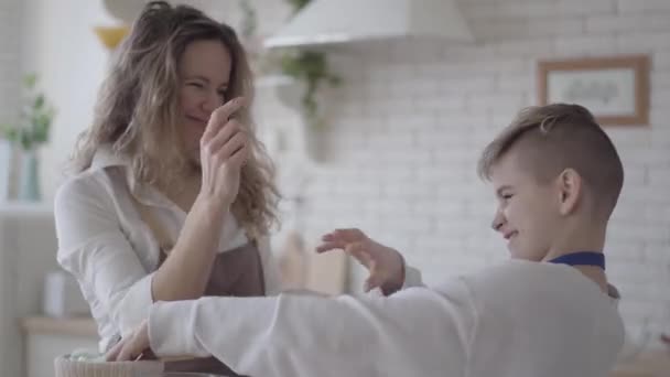 Mulher bonita e seu filho adolescente cozinhar bolo na cozinha, o menino ajudando a mãe a fazer comida. Mamãe tentando manchar o rosto do garoto com farinha, ambas as pessoas sorrindo. Família feliz se divertir em casa — Vídeo de Stock