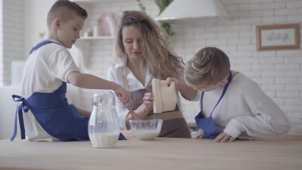 Pretty kvinna och hennes två söner matlagning göra degen i köket, pojkarna hjälper mamma att göra mat. Mamma hälla mjöl i skålen och Kid blanda det. Glad vänlig familj — Stockvideo