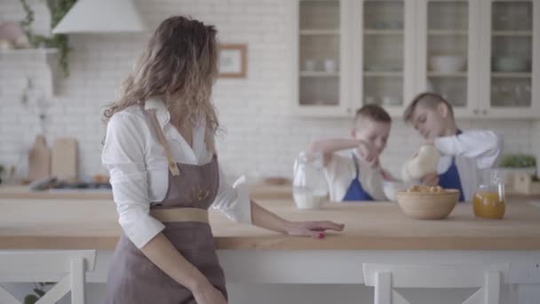 Mulher bonita de avental em primeiro plano olhando para seus dois filhos cozinhar fazendo massa na cozinha com sorriso feliz. Os rapazes a ajudarem a mãe a fazer comida. Família amigável feliz — Vídeo de Stock