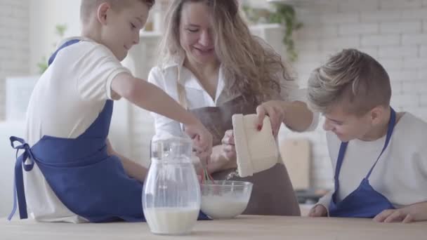 Portrait pretty woman and her two sons cooking making dough in the kitchen, the boys helping mother to make food. Mom pouring flour in the bowl and kid mixing it. Happy friendly family — Stock Video