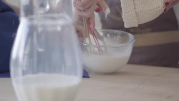 Close up view cooking making dough for baking pie, cake, bread. Hands of unrecognizable woman pour flour in the bowl and mixing it with a whisk. Jug of milk is in the foreground on the table — Stock Video