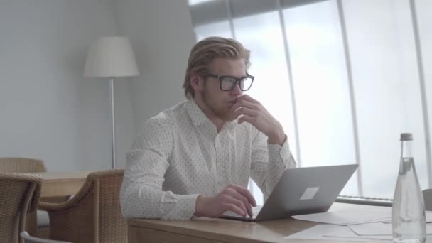 Блондинка вдумлива людина в окулярах сидить за столом у легкому комфортному офісі перед нетбуком. Красивий бізнесмен погладжує свою бороду, думаючи про роботу . — стокове відео