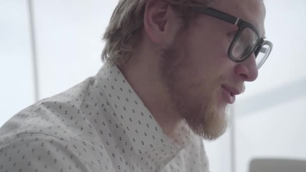 Cerca de cara barbudo hombre de negocios en gafas sentado en una oficina cómoda luz. Un hombre de negocios guapo trabajando. Vista lateral. Lugar de trabajo . — Vídeos de Stock