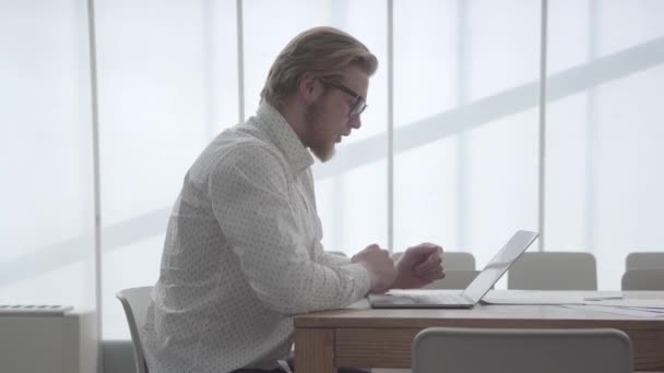 Retrato guapo rubio pensativo hombre de negocios en gafas sentado a la mesa en una oficina cómoda luz delante de netbook . — Vídeos de Stock