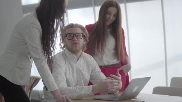 メガネをかけたブロンドの男は、同僚に自分の考えを説明するテーブルのネットブックを持つ明るい快適なオフィスに座っています。上司の話を聞いて近くに立っているフォーマルな服を着た2人の女の子. — ストック動画