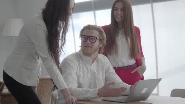 Retrato exitoso hombre rubio en gafas sentado en una oficina cómoda luz con netbook en la mesa explicando su idea a los colegas. Dos chicas en ropa formal de pie cerca de escuchar al jefe . — Vídeos de Stock