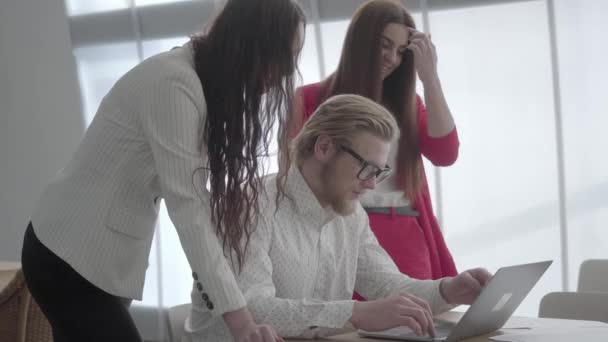 Blond man i glasögon sitter i en ljus bekväm kontor med Netbook vid bordet förklarar sin idé till kollegor. Två flickor i formella slitage stående nära lyssnar på Boss. — Stockvideo