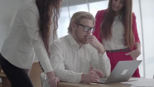 Porträtt framgångsrik blond man i glasögon sitter i en ljus bekväm kontor med Netbook vid bordet förklarar sin idé till kollegor. Två flickor i formella slitage stående nära lyssnar på Boss. — Stockvideo