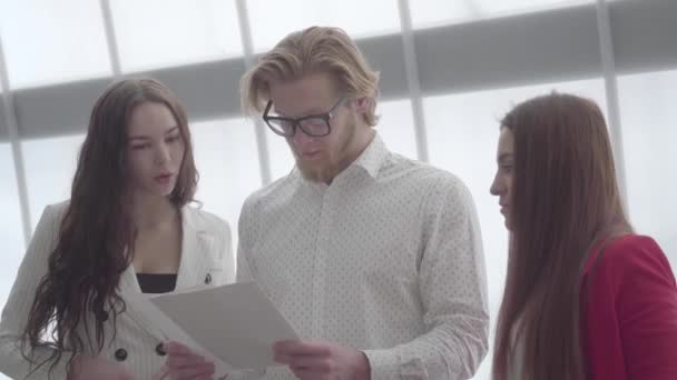 Retrato exitoso hombre rubio en gafas explicando su idea a dos colegas femeninos de pie a cada lado de él en una oficina cómoda y ligera. Dos chicas en ropa formal escuchando al jefe — Vídeo de stock