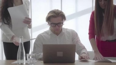 Portre başarılı sarışın adam gözlük lüzumlu rahat bir ofiste oturan netbook ile masada imza belgeleri meslektaşları ona getirdi. Ya ayakta resmi giyim iki kız