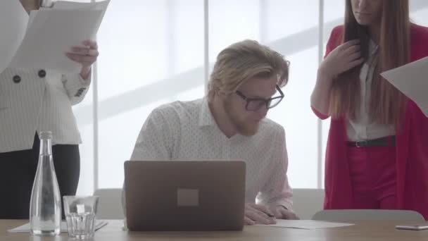 Portret pomyślny biznesmen w okularach siedzi w jasnym biurze wygodne z Netbook przy stole podpisywania dokumentów, które koledzy przyniósł do niego. Dwie dziewczyny w odzieży formalnej — Wideo stockowe